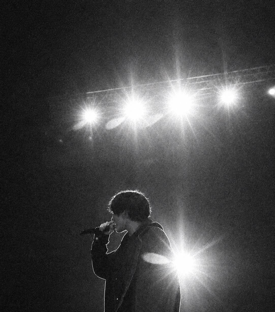 Phil Coutier in profile, singing into a microphone beneath bright stage lights.