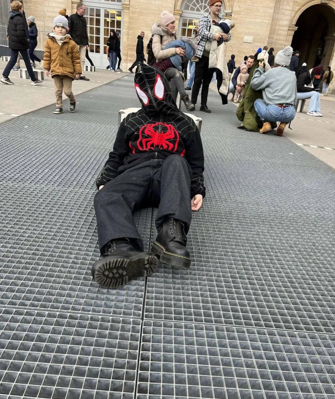 A seated figure in a black and red Spider-Man hoodie in a busy public square.